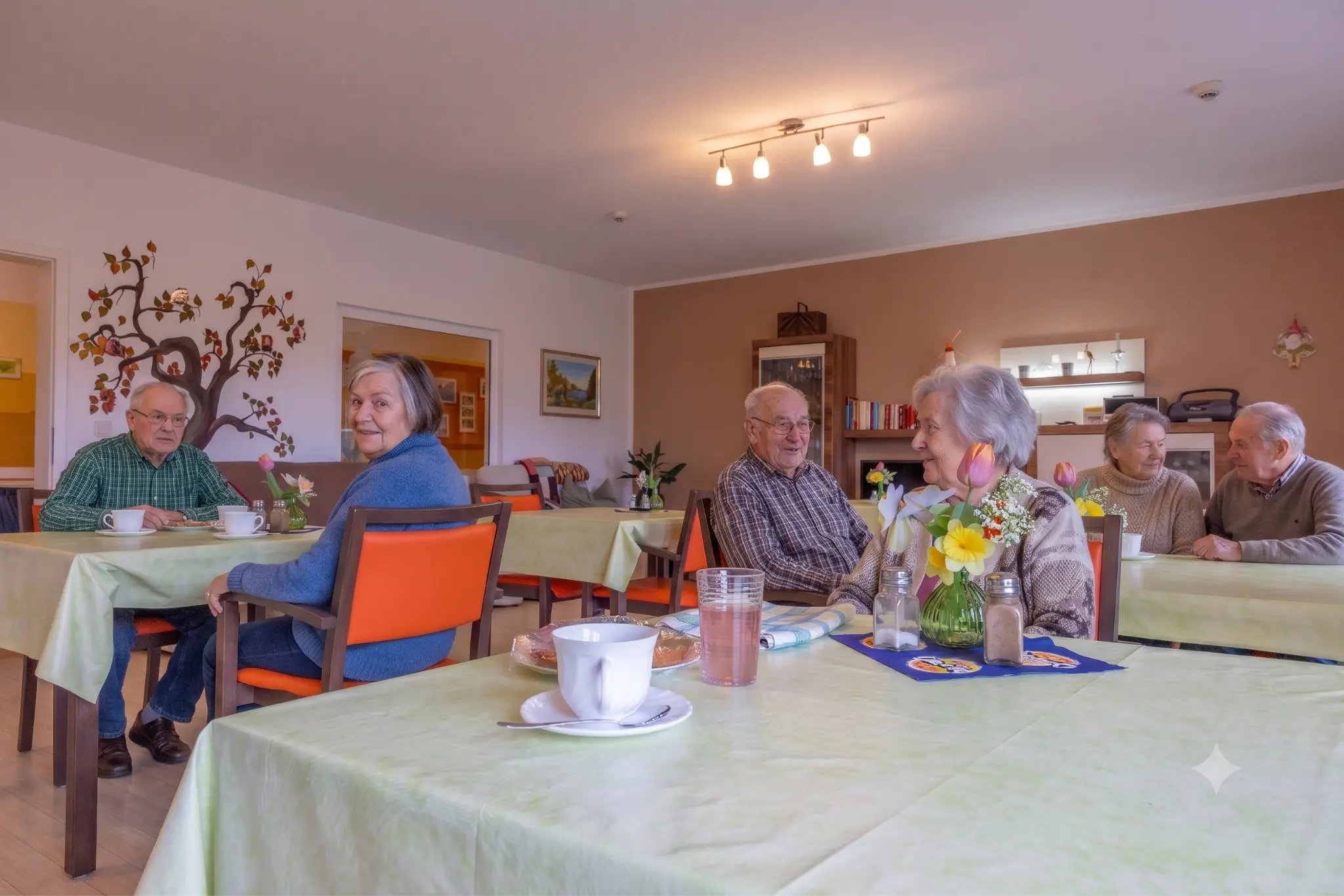 Mehrere Bewohnerinnen und Bewohner der Senioren-WG sitzen gemeinsam an Tischen im Aufenthaltsraum und trinken Kaffee.