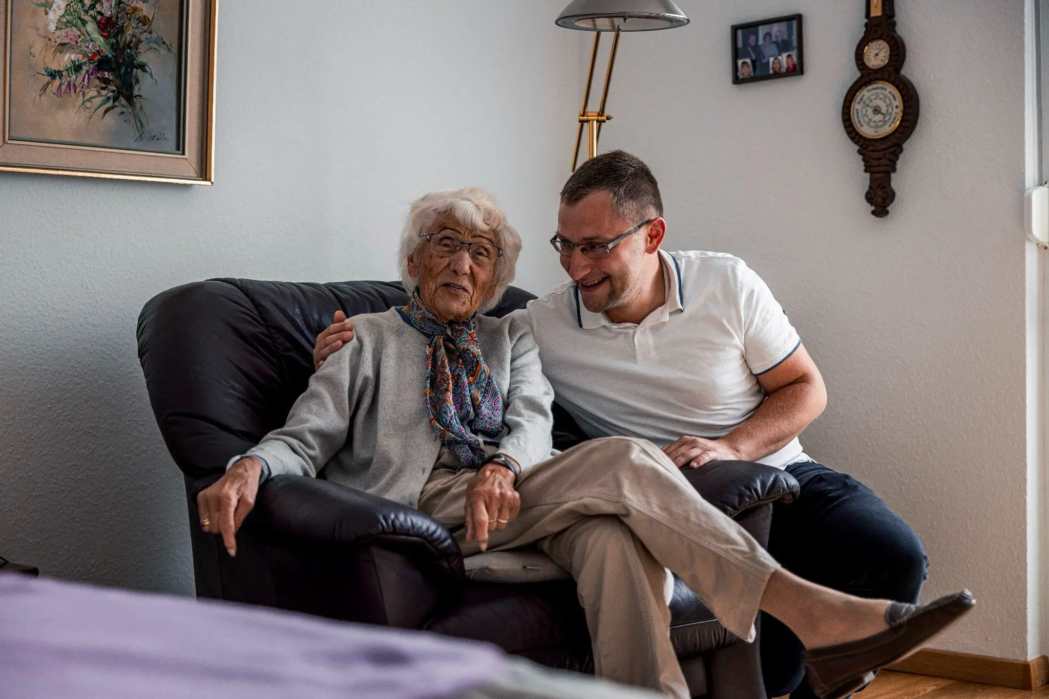 Ein Mitarbeiter des Pflege- und Betreuungsdienstes Müller sitzt neben einer Bewohnerin auf dem Sofa, hat den Arm um sie gelegt, beide lächeln.