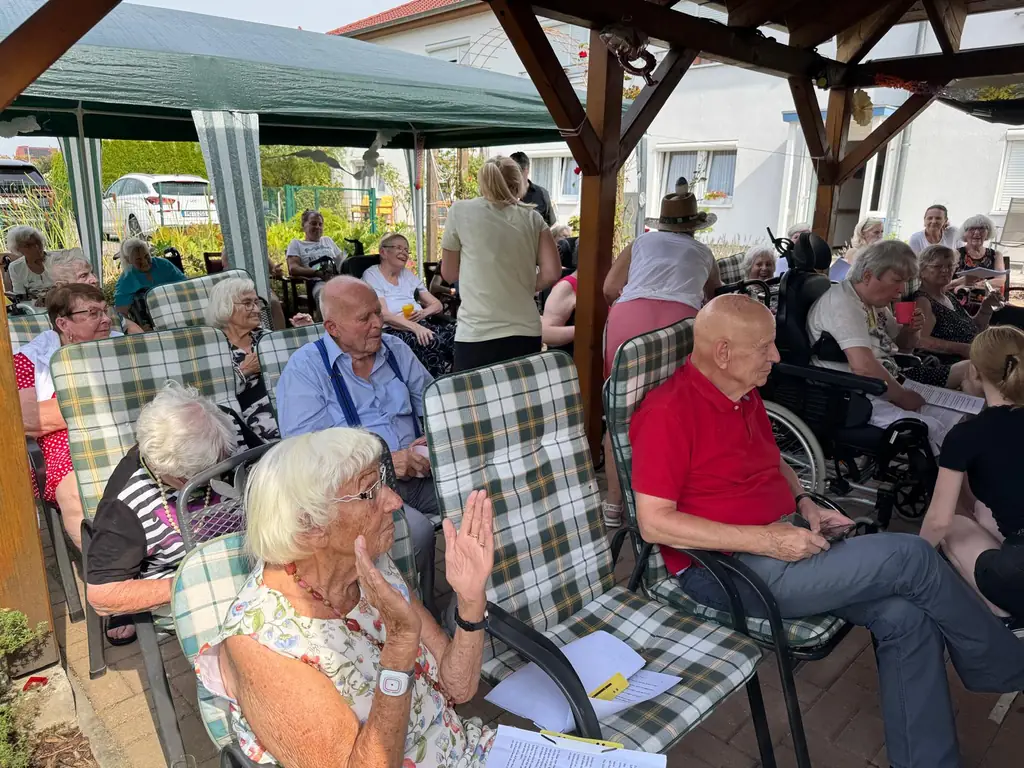 Bewohnerinnen und Bewohner der Senioren-WG des Pflege- und Betreuungsdienstes Müller sitzen bei einem Sommerfest gemeinsam unter der überdachten Pergola im Garten.